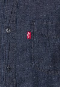 Mörkblå denimskjorta med ficka, svarta knappar och en röd Levi's-lapp. Texturen är något grov med en slät finish.