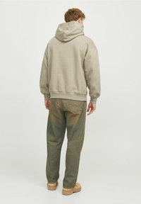 Sudadera beige clara con capucha grande, corte holgado y puños acanalados. Combinada con pantalones vaqueros verdes desgastados y calzado beis. Vista trasera, fondo liso.