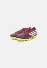Zapatos de fútbol en color marrón con parte superior de sintético texturizado, marca de tres rayas en blanco y tacos amarillos brillantes para tracción.
