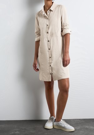 Femme portant une robe beige boutonnée avec des manches retroussées et des baskets blanches, debout contre un mur blanc uni et un sol sombre.