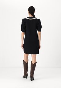 Zwarte korte jurk met pofmouwen, voorzien van een witte decoratieve kraag. Model draagt bruine cowboylaarzen met puntige toppen. Effen achtergrond.