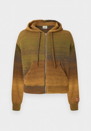 Junger Mann trägt grünen und braunen Farbverlauf-Hoodie, weißes Grafik-T-Shirt, locker sitzende hellblaue Jeans, schwarzen Gürtel und braune Stiefel, steht mit einer Hand in der Tasche.