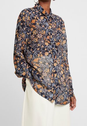 Femme portant un chemisier transparent à manches longues avec un motif floral multicolore et un pantalon blanc taille haute.