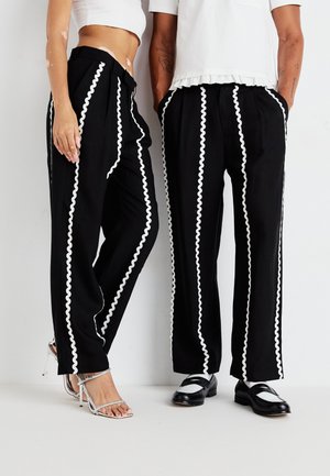 Black pants with white wavy contrast stripes, featuring a relaxed fit and pleats. One pair worn with silver heels, the other with black shoes.