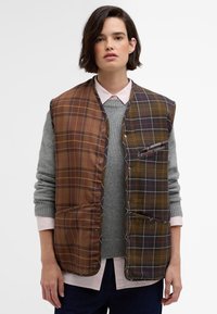 Hnědá a zelená kostkovaná vesta s patentkami, která má dvě přední kapsy, nošena přes šedý svetr a růžovou košilí s límečkem. Hladká textura.