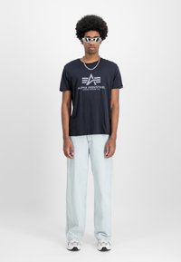 Marineblå bomulds t-shirt med "Alpha Industries" logo, lyseblå denim jeans, sølvfarvede briller og sneakers. Modellen står foran en ensfarvet baggrund.