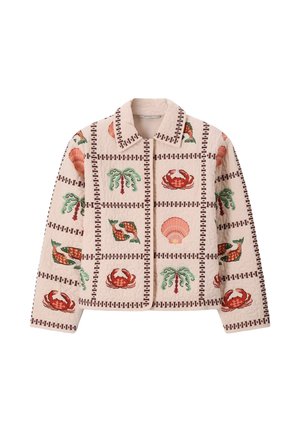Femme portant une veste à motifs et un pantalon à jambes larges, tenant un grand sac cabas fleuri, debout devant un fond beige.
