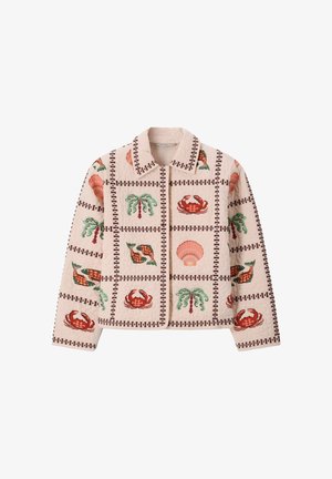 Vrouw draagt een jas met patroon en broek met wijde pijpen, houdt een grote bloemige tas vast, staand tegen een beige achtergrond.