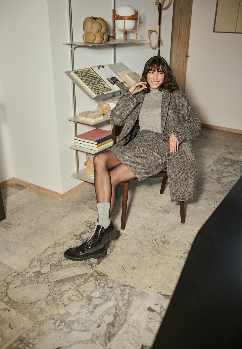 Grey plaid blazer over a grey turtleneck, paired with a matching skirt. Black lace-up shoes and grey socks. Room features modern decor and books.