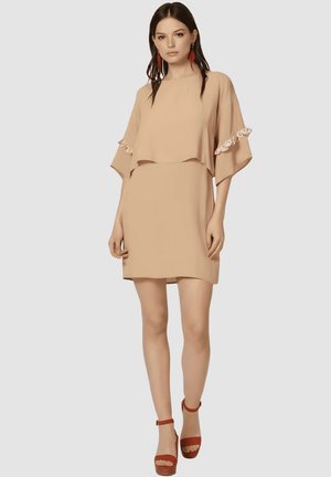 Mujer con un vestido corto beige de mangas anchas y detalles florales, combinado con sandalias de plataforma rojas y pendientes de borlas rojas.