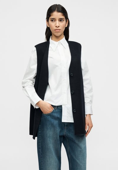 Μαύρο αμάνικο γιλέκο με κουμπιά στο μπροστινό μέρος, φορεμένο πάνω από λευκό πουκάμισο με γιακά και μπλε τζιν. Το γιλέκο έχει ομαλή υφή και κομψό σχέδιο.