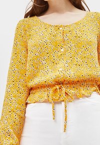 Gul blus med blommönster, långa ärmar, rund halsringning, knäppning fram och en rynkad nederkant med snöre. Har ett prästkrage-mönster och är tillverkad av lättviktsmaterial.