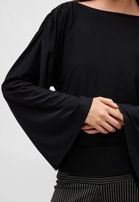 Must pikkade varrukatega pluus, millel on lai kaelus, õlgadel kogutud detailid ja lehvitatud varrukad, mida on kombineeritud istuvate triibuliste pükstega.