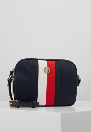 Laivastonsininen olkalaukku, jossa punaisia, valkoisia ja mustia pystysuoria raitoja sekä kultainen Tommy Hilfiger -logo edessä, asetettuna valkoiselle pinnalle.