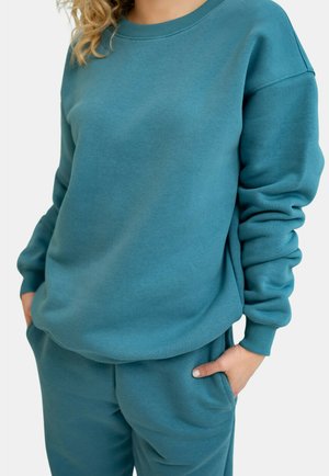 Osoba ubrana w morski, długi rękaw bluzy i dopasowane spodnie, z rękami w kieszeniach, prezentująca swobodny, casualowy strój.