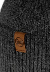 Gestrickte graue Mütze mit gerippter Textur und einem braunen Lederetikett mit dem "Buff"-Logo. Nahaufnahme des Stoffes.