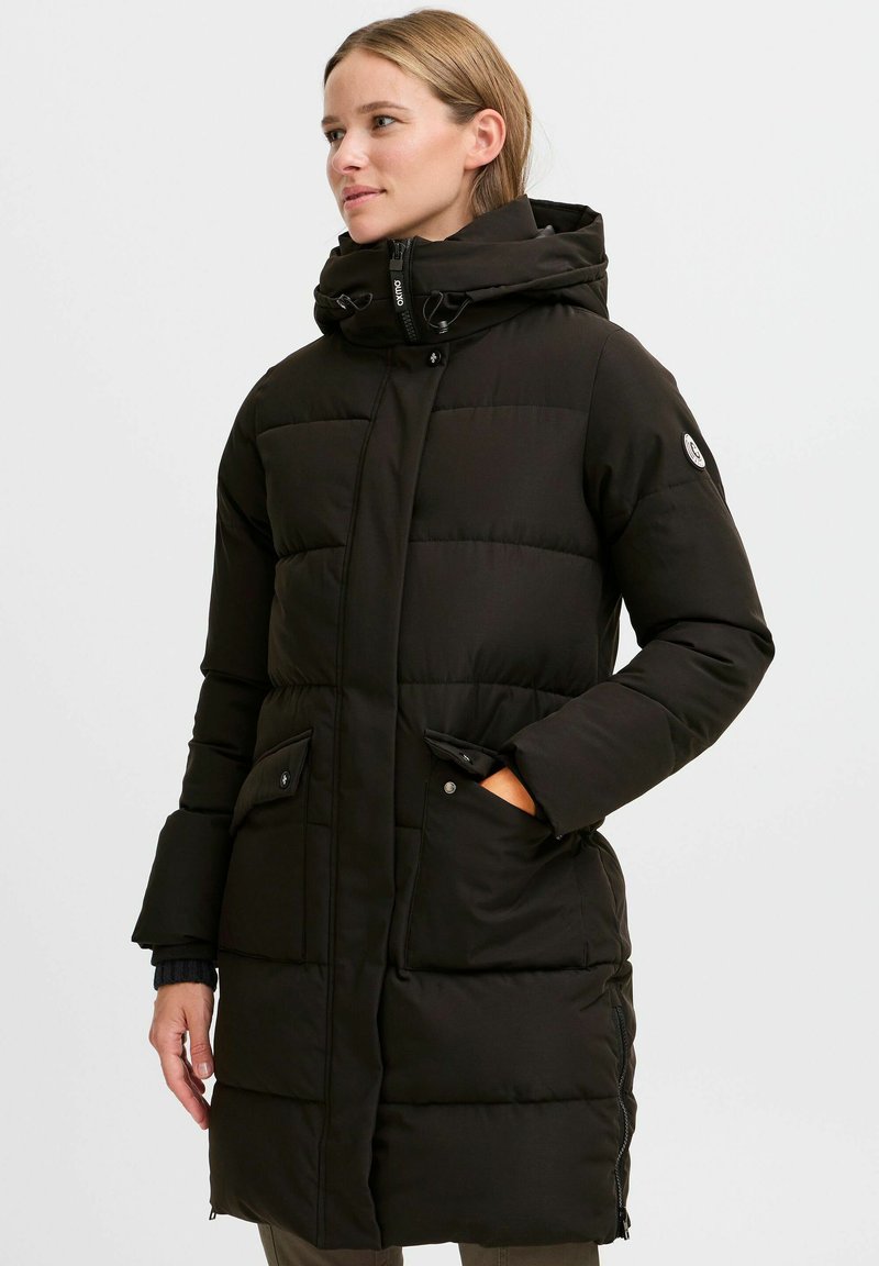 Black puffer jacket with a high collar, quilted design, front zip closure, and two snap-button pockets. Features a logo patch on the arm.