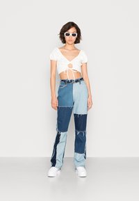 Top branco curto com mangas bufantes e detalhe de laço na parte da frente. Calças de ganga azul com remendos, apresentando tons variados e bordas desfiadas. Sapatos brancos.