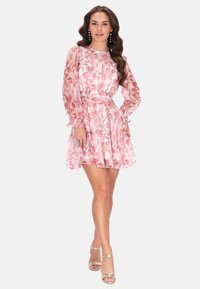 Różowa sukienka w kwiaty z długimi przezroczystymi rękawami, talią podkreśloną paskiem i falbaną na dole, zestawiona z srebrnymi sandałami na paski. Gładki, lekki materiał.