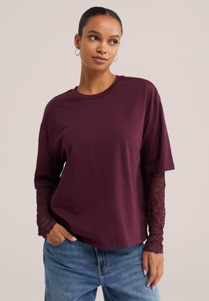 Femme portant un t-shirt ample violet foncé par-dessus un haut en dentelle violet à manches longues et un jean bleu, une main dans la poche et regardant sur le côté.