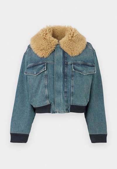 Džinsinė striukė su dideliu smėlio spalvos dirbtinio kailio apykakle, priekinėmis kišenėmis su sklendėmis, ribinėmis rankogaliais ir ribintu liemeniu ant balto fono.