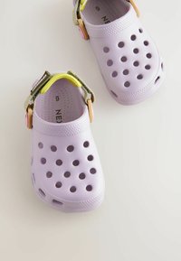 Zoccoli di un colore viola chiaro con fori di ventilazione e una cinghia con un accento verde. Realizzati in materiale simile alla gomma, superficie testurizzata.