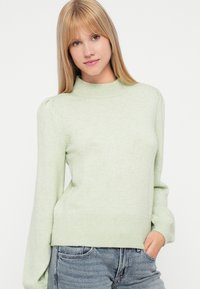Gaiši zaļš adīts džemperis ar augstu apkakli, puffed pleciem, ribotu apakšmalu un apkaklēm, apvienots ar ziliem džinsu biksēm. Mīksta tekstūra un pieguļošs dizains.