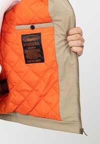 Pikowana beżowa kurtka bomberka z pomarańczową podszewką, z kieszonką zapinaną na guzik i naszywką z logo, trzymana w ręce.