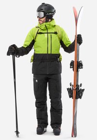 Équipement de ski comprenant une veste imperméable verte fluo et noire, un pantalon de ski noir, des gants, un casque et des skis avec des fixations noires et des accents oranges.