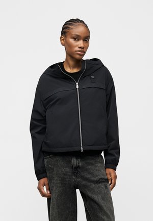 Młoda kobieta z zaplecionymi włosami, ubrana w czarną kurtkę z kapturem na zamek oraz wyblakłe czarne jeansy, na jasnym, prostym tle.