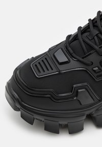 Nahaufnahme eines schwarzen Sneakers mit dicker, robuster Sohle, strukturiertem Obermaterial, verstärkter Zehenpartie und schwarzen Schnürsenkeln auf weißem Hintergrund.