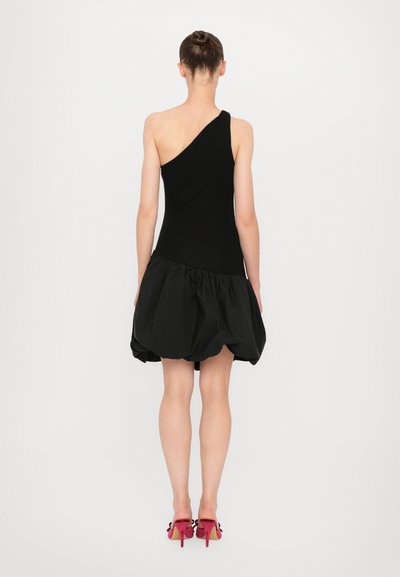 Robe noire à une épaule avec un corsage ajusté et une jupe volumineuse texturée. Portée avec des chaussures à talons hauts roses ornées de nœuds.