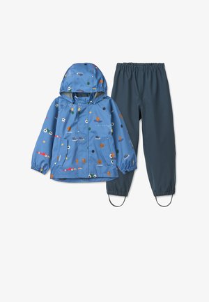 Giacca impermeabile blu con cappuccio, caratterizzata da stampe colorate a tema sportivo, abbinata a pantaloni grigio scuro con vita ed elastici alla caviglia.