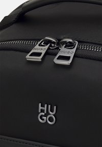 Sac en tissu noir avec deux tirettes de fermeture éclair argentées et logo "HUGO" argenté sur le panneau avant.