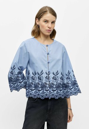 Femme portant un chemisier bleu clair avec broderie florale bleu marine et bordure en dentelle, associée à un pantalon noir, debout sur fond blanc.