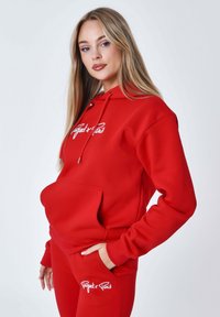 Felpa rossa con cappuccio e pantaloni abbinati, realizzati in tessuto morbido, con tasca frontale e logo bianco. Il design include lacci nel cappuccio.