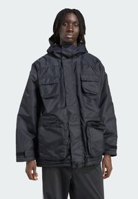 Person med mørk hud og dreadlocks iført en sort hættetrøje med fire store frontlommer, stående mod en ensfarvet lys baggrund.
