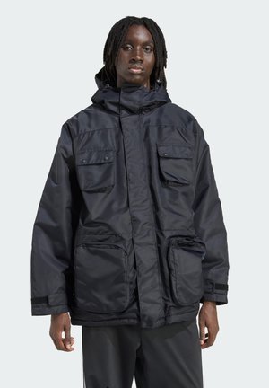 Person with dark skin and dreadlocks wearing a black hooded jacket with four large front pockets, standing against a plain light background.