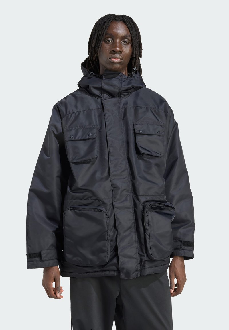 Person med mørk hud og dreadlocks iført en sort hættetrøje med fire store frontlommer, stående mod en ensfarvet lys baggrund.