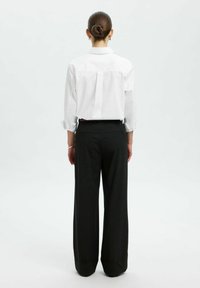 Witte blouse met lange mouwen en een rugpas, gecombineerd met zwarte ruime broek. Model gezien van achteren, haar in een lage knot.