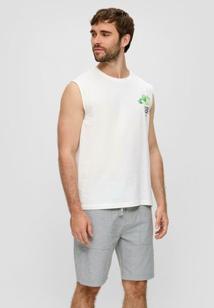 Weißes ärmelloses Baumwollshirt mit einem grünen Grafikdruck auf der Brust, kombiniert mit hellgrauen gemusterten Shorts.