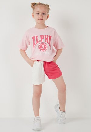 Niña joven con el cabello trenzado, vestida con una camiseta rosa que dice "Alpha Girl Team", pantalones cortos bicolor rojo y blanco, y zapatillas blancas, posando con confianza.