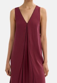Bordowa sukienka bez rękawów z dekoltem w kształcie litery V, z czarnymi przeszyciami wzdłuż dekoltu oraz z plisowanym wzorem, który dodaje tekstury.