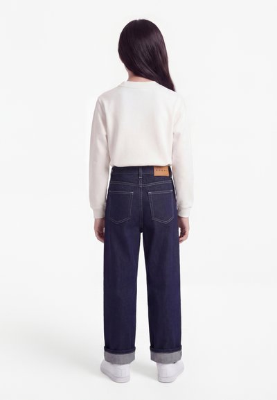 Παιδί με μακριά σκουρόχρωμα μαλλιά στέκεται γυρισμένο προς τα πίσω, φοράει λευκό μακρυμάνικο πουκάμισο, σκούρο μπλε τζιν με μανσέτες και λευκά αθλητικά παπούτσια.