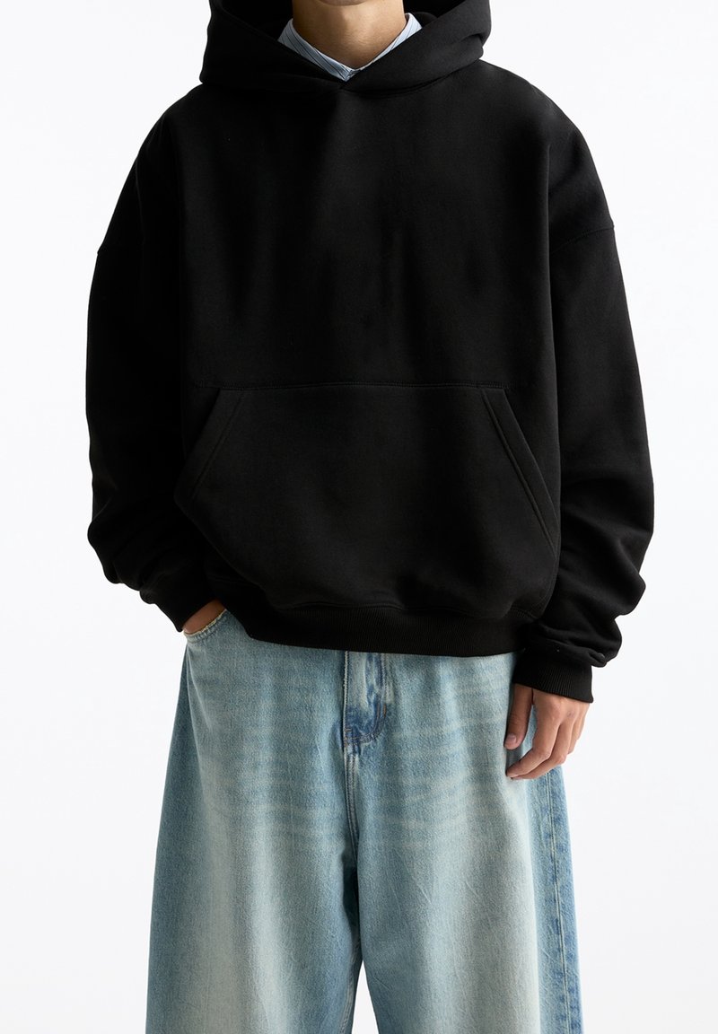 Personne portant un sweat à capuche noir avec une poche avant et un jean large bleu clair, debout avec une main dans la poche.