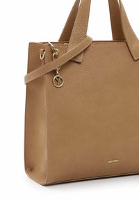 Tote-Bag aus tanfarbenem Leder mit strukturierter Oberfläche, ausgestattet mit doppelten Griffen, einem abnehmbaren Riemen und einem goldenen Charme als Akzent. Markenname sichtbar.