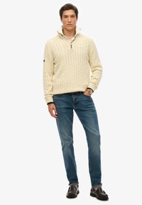 Crème kabelgebreide pullover met rits, gedragen over een wit shirt. Gestyled met blauwe jeans en donkerbruine instappers, wat een casual look laat zien.