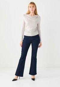 Jasnoszara bluzka z długim rękawem i drapowanym dekoltem, zestawiona z ciemnoniebieskimi rozszerzanymi dżinsami oraz czarnymi szpilkami. Gładki materiał o miękkiej fakturze.