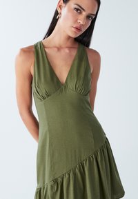 Olivgrünes Kleid mit V-Ausschnitt, ärmellosem Design, gerafftem Oberteil und gerüschtem Saum, gefertigt aus weichem Stoff mit glatter Textur.