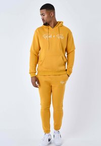 Sudadera con capucha amarilla con logo blanco, bolsillo frontal y cordones, combinada con joggers cónicos a juego. Zapatillas blancas completan el conjunto.